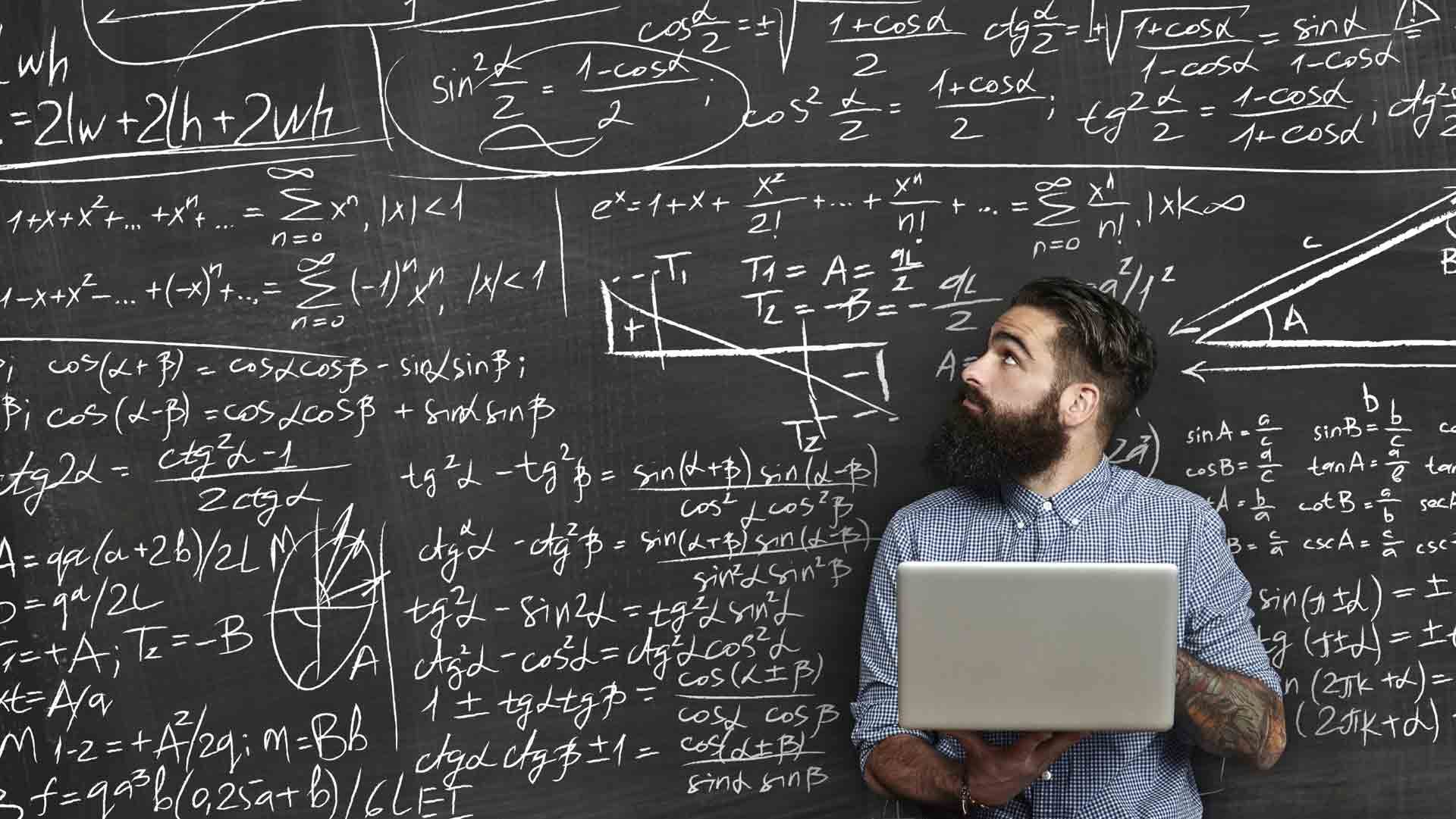 Преподаватель с ноутбуком у школьной доски, исписанной математическими формулами